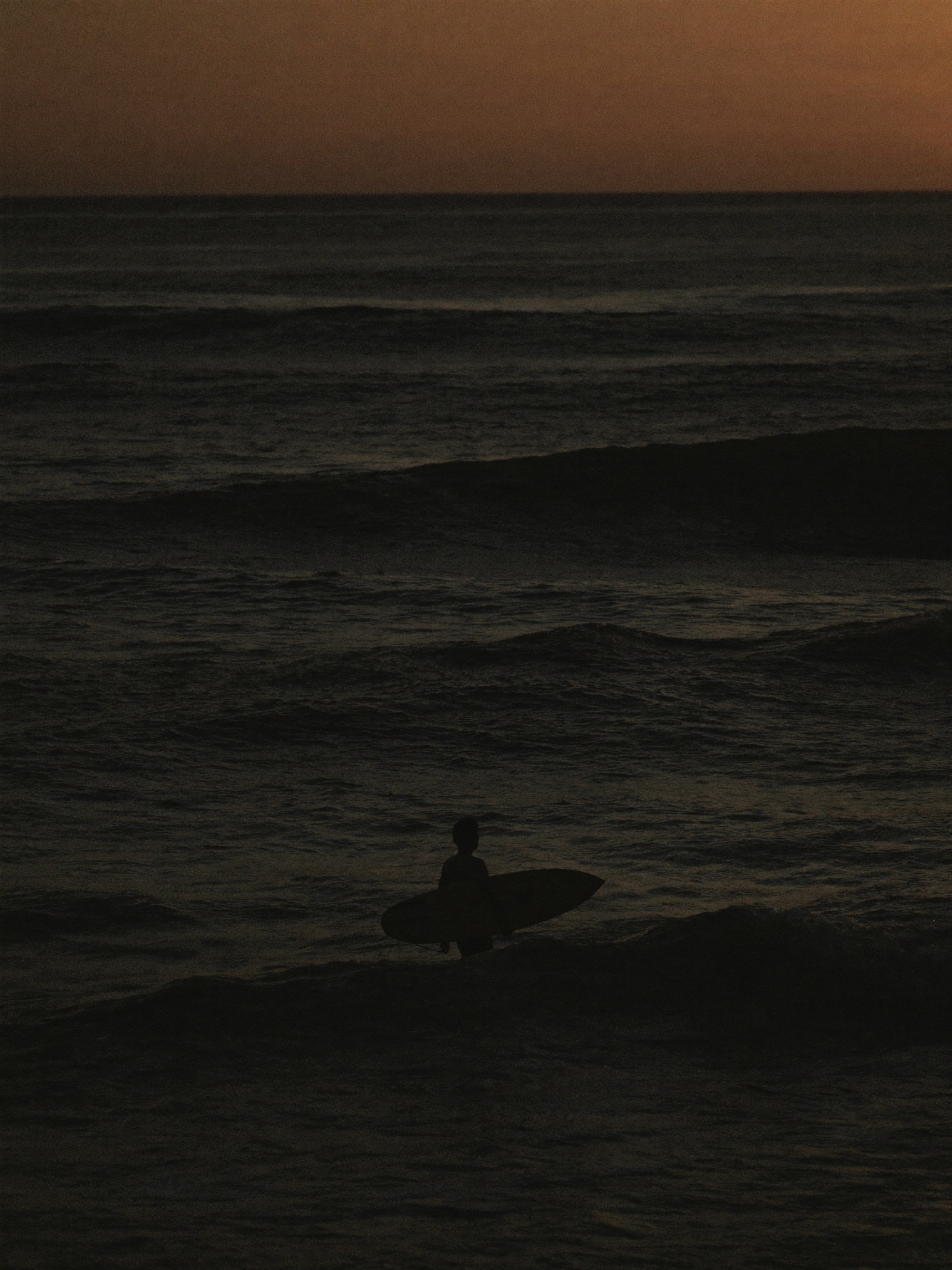 Surfer walking into a dark ocean at dusk